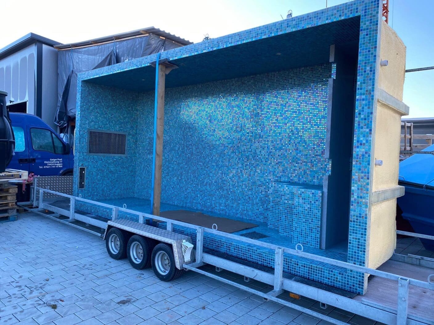 Blue-tiled pool shell rests on a triple-axle trailer, empty and supported by a vertical wooden post in a paved industrial yard beside vehicles and pallets. Text on van (partially legible): "Geiger Pool" "Tel. 0761 / 93 16 900" (other small text illegible).