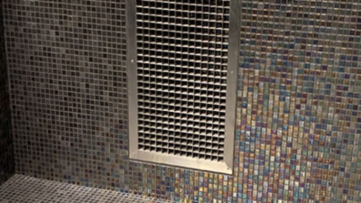 Rectangular metal vent grille is mounted on a bathroom wall covering an air opening, surrounded by iridescent square mosaic tiles that continue onto the shower floor and a recessed niche.