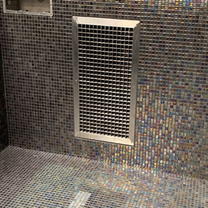 Rectangular metal vent grille is mounted on a bathroom wall covering an air opening, surrounded by iridescent square mosaic tiles that continue onto the shower floor and a recessed niche.