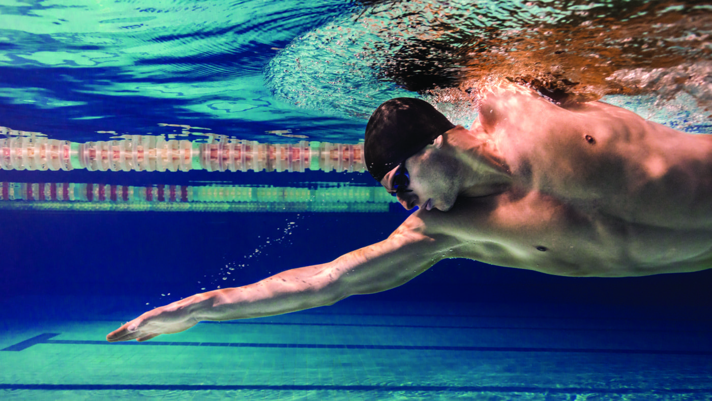 A swimmer in a black swim cap and goggles extends his arm in a streamlined freestyle stroke underwater alongside a lane divider in a clear blue pool.