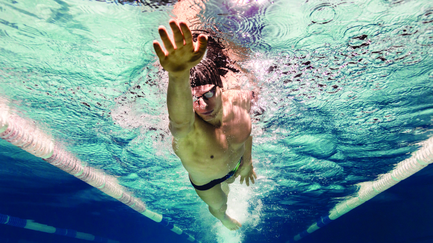 Competitive swimmer reaches forward mid stroke while propelling through turquoise lane of a pool, wearing goggles and cap, bubbles and rippling surface visible between white lane dividers.