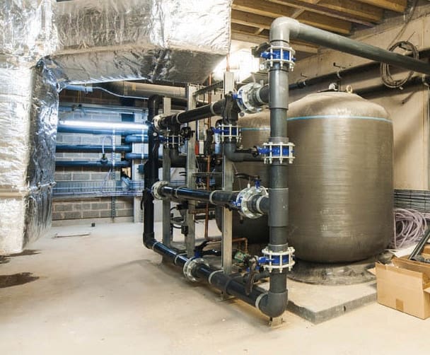 A black multi tier pipe assembly connects to a large vertical water tank, running across a concrete floored mechanical room with foil insulated ducts, exposed ceiling joists and a box.