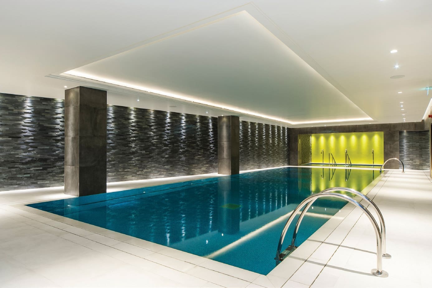Indoor swimming pool sits still, reflecting recessed ceiling lights; surrounded by white tile deck, dark textured stone walls with pillars, and a yellow-lit alcove at the far end.