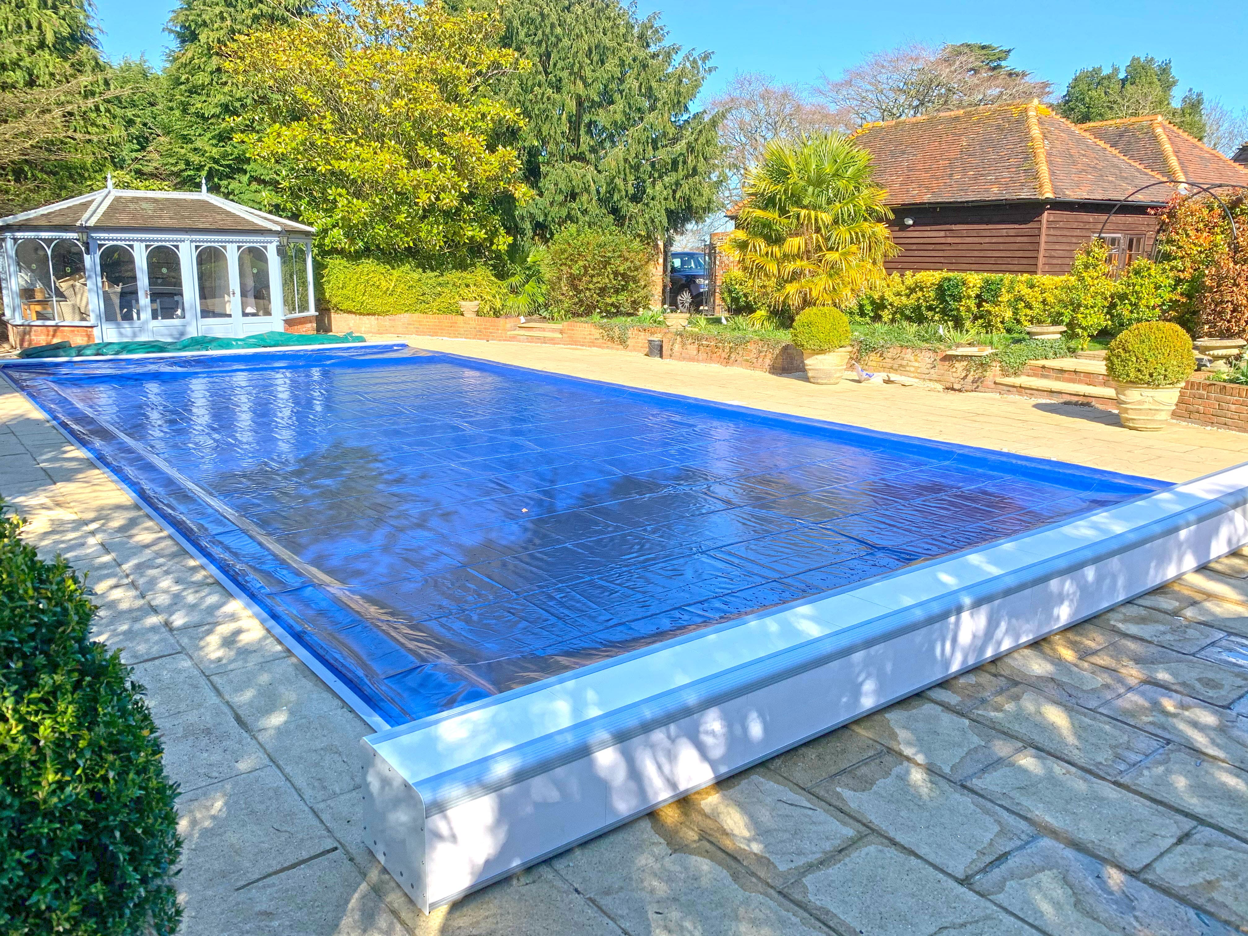 A rectangular pool covered by a taut blue solar cover sits on a stone patio beside a white gazebo, flanked by potted shrubs, hedges, trees and a wooden outbuilding.