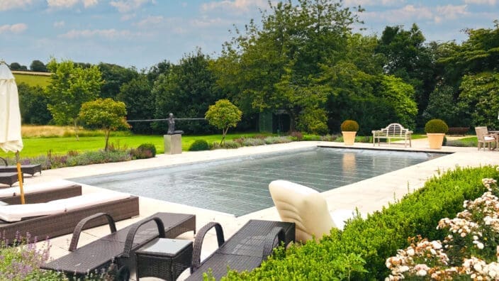 A rectangular pool with a dark cover sits among patio loungers and flowerbeds, flanked by potted topiaries and benches, set before an expansive lawn and treeline under a blue sky.