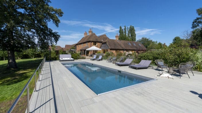 Rectangular swimming pool reflects blue sky, bordered by pale deck; loungers and a spotted dog lie beside it, with a large brick house and trees in the sunny garden.