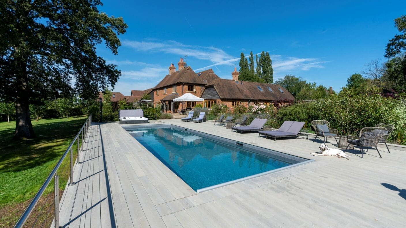 Rectangular swimming pool sits within a wide pale wood deck, reflecting the blue sky; lounge chairs and a Dalmatian recline nearby, with a large brick house and trees in the background.