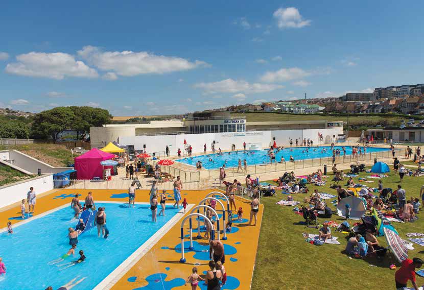 Outdoor lido SALTDEAN LIDO with families swimming and children playing in splash pools while others sunbathe on a grassy terrace under a clear blue sky beside a seaside town.