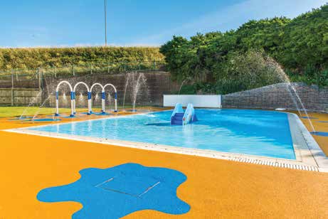 Children's splash pool with a blue slide and arched water jets spraying over it, on a bright orange deck with blue splash motif, backed by grassy berm and stone wall.