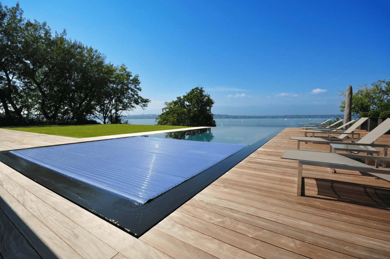 Infinity pool reflecting sky sits partially covered by a blue automatic cover, flanked by wooden deck with lounge chairs, overlooking a calm lake and tree-lined shore under clear blue sky.