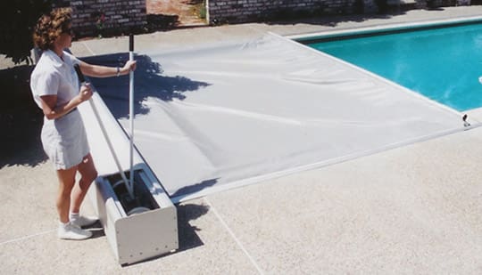 Pool cover being pulled across a residential swimming pool while a woman operates a manual reel on the concrete pool deck.