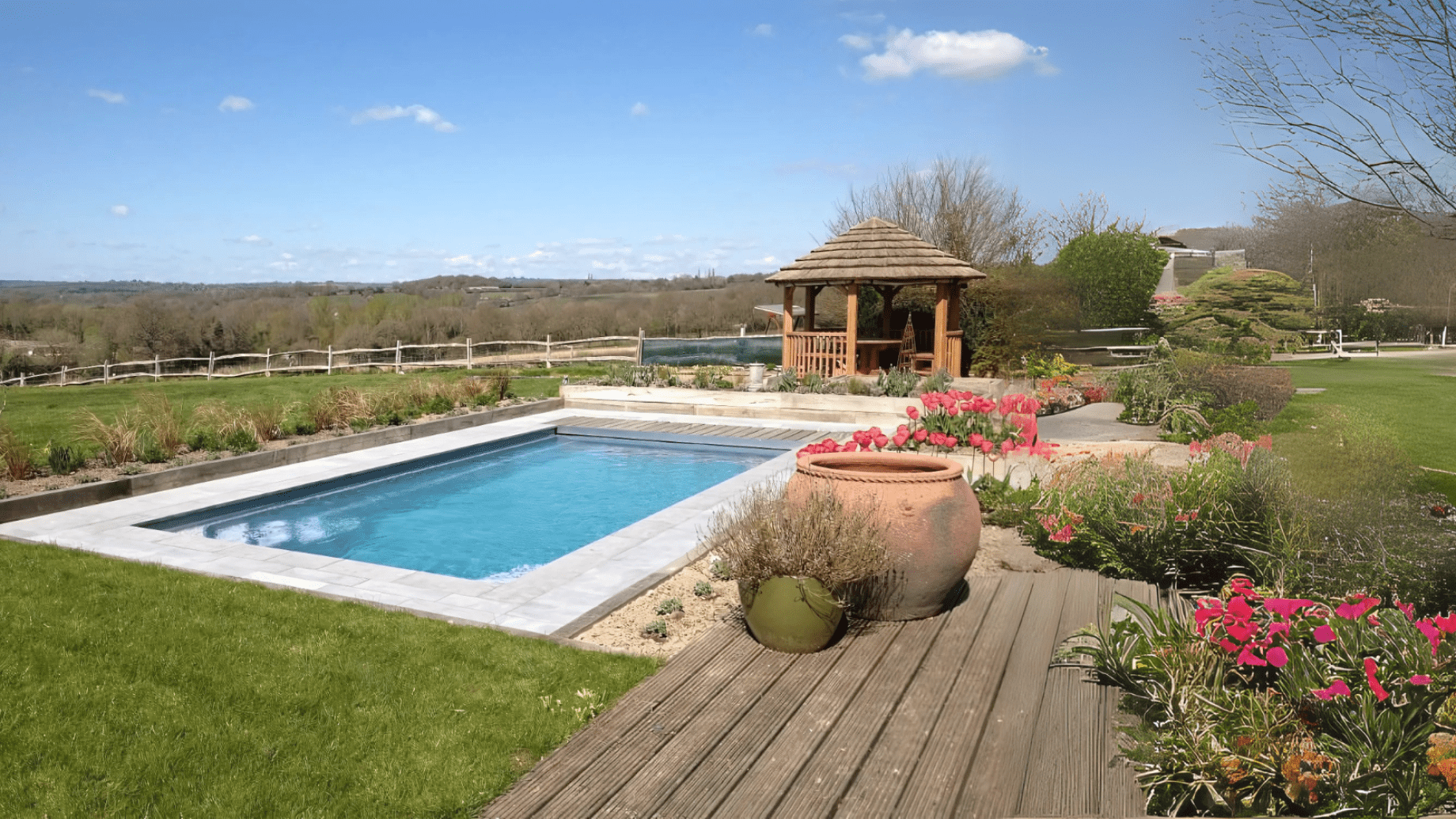 Rectangular blue swimming pool sits amid wooden decking and flowerbeds, with terracotta pots and pink tulips nearby, a wooden gazebo behind, and rolling countryside under a clear blue sky.