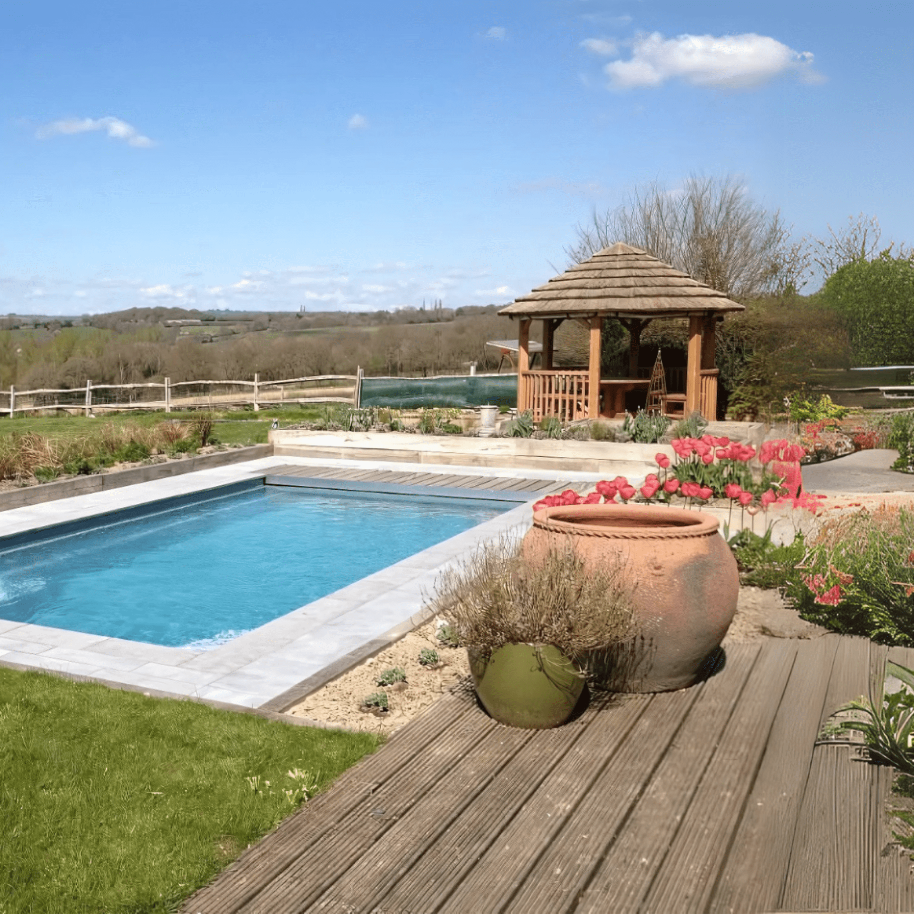 Rectangular blue swimming pool sits amid wooden decking and flowerbeds, with terracotta pots and pink tulips nearby, a wooden gazebo behind, and rolling countryside under a clear blue sky.