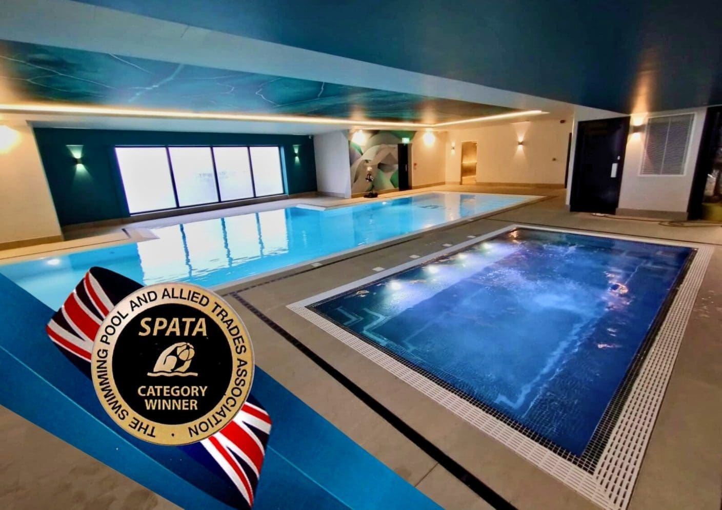 Indoor rectangular swimming pool and adjacent bubbling spa reflecting ceiling lights, set in a modern tiled leisure room with large frosted windows and wall sconces; Badge text: SPATA CATEGORY WINNER THE SWIMMING POOL AND ALLIED TRADES ASSOCIATION