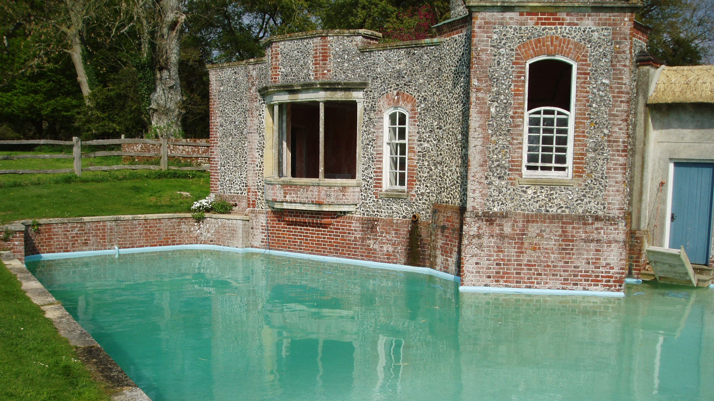 Stone and brick building faces a turquoise reflecting pool, windows open and partially boarded, a pipe drains water into the pool, grassy lawn and wooden fence surround the scene